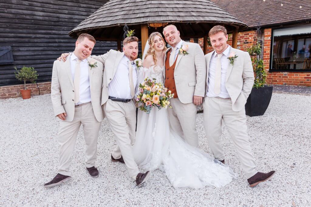 A bride in a white gown stands with four groomsmen in light suits and ties, posing playfully outside a rustic wooden gazebo. The perfect wedding photographer captures their warm smiles as the bride holds a colorful bouquet of flowers.