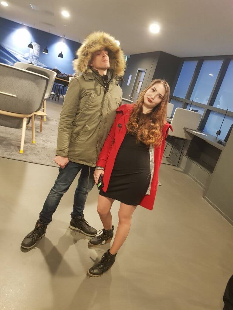 A person in a green parka with a fur hood stands playfully next to another in a red coat and black dress. The modern, indoor setting with large windows creates the perfect wedding photo opportunity, capturing their lighthearted, casual pose effortlessly.