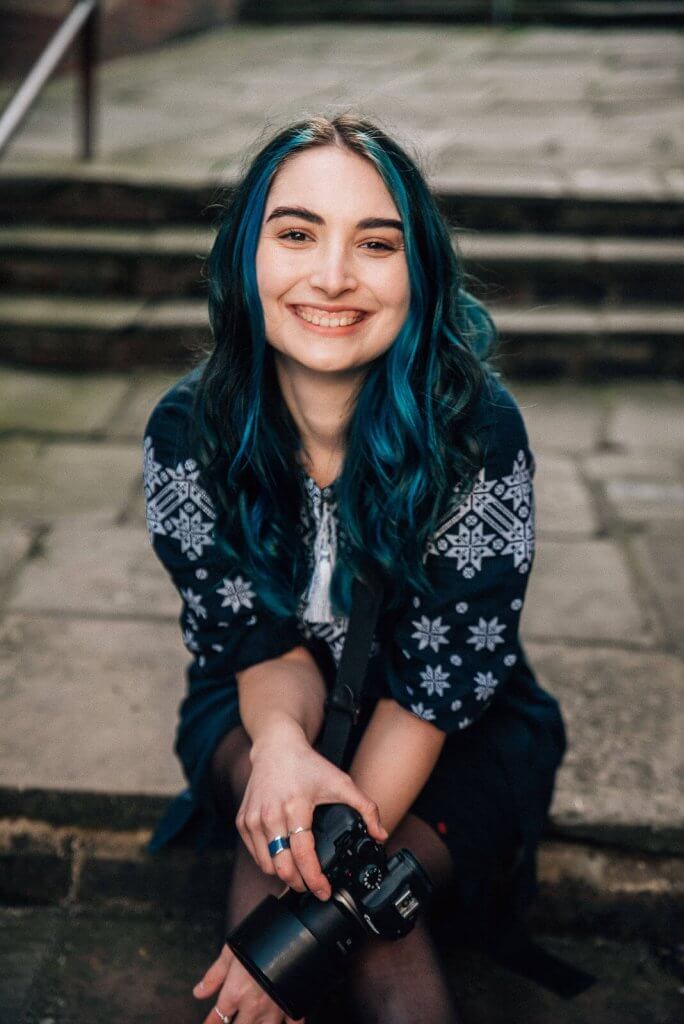 A person with long blue hair, looking for the perfect wedding photographer, smiles at the camera while sitting outdoors on stone steps. They wear a dark patterned sweater and hold a camera in their hand. The setting appears casual and relaxed.