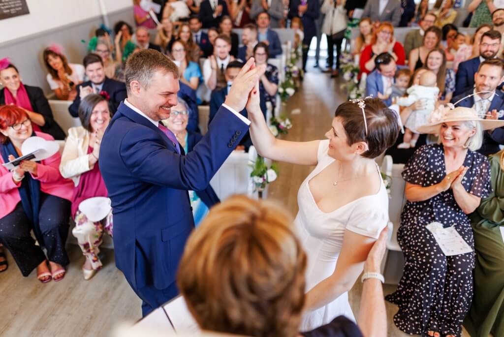 A couple in wedding attire high-five during their ceremony, surrounded by an applauding crowd of guests in a bright room. The groom is in a blue suit and the bride in a white dress, both smiling. The scene, captured by the perfect wedding photographer, includes flowers and greenery.