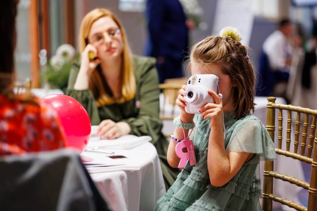 A young girl in a green dress sits at a table, snapping a photo with her white instant camera. A pink balloon graces the table, while in the background, a woman with glasses watches thoughtfully—a scene capturing life's moments as skillfully as the perfect wedding photographer would.