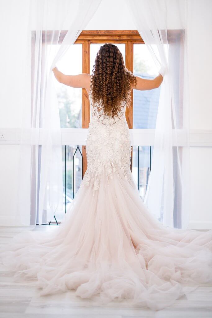 A bride in a lace wedding gown with a long train stands facing a window, holding the sheer curtains open. Her curly hair cascades down her back as she looks outside, creating a serene and elegant scene—capturing the essence of a perfect wedding.