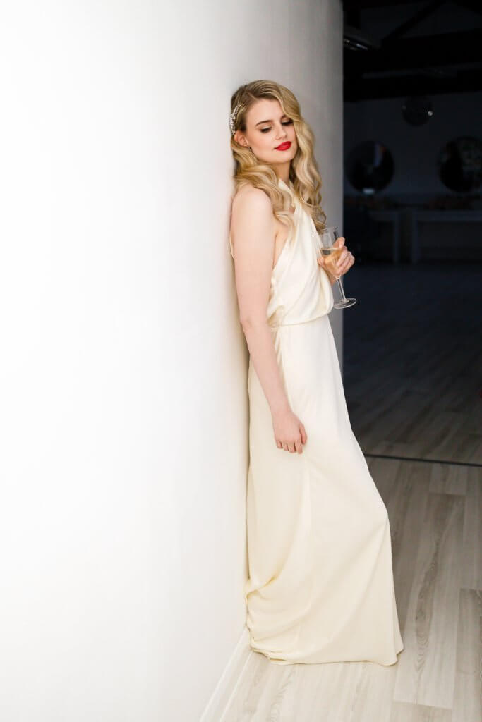 A woman in an elegant white gown leans against a white wall, wine glass in hand. With long, wavy blonde hair and red lipstick, she exudes timeless romance—ideal for capturing by the perfect wedding photographer under the room's dim lighting and against the rich wooden floor.
