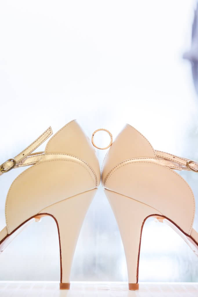 A pair of cream-colored high-heeled shoes face each other, with a gold ring balanced between their heels. The background is softly blurred, highlighting the elegance of this perfect wedding moment.