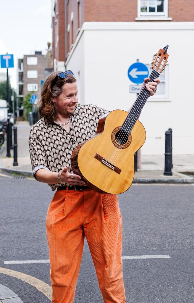 A person with long hair stands on a city street, acoustic guitar in hand. They're dressed in an abstract patterned shirt and orange pants, sunglasses perched on their head—a true artist's vibe. Perhaps they're searching for the perfect wedding photographer amid the backdrop of urban life and distant traffic signs.