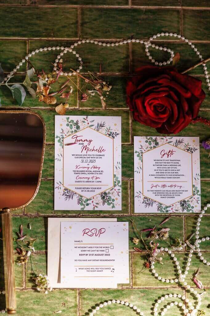 Wedding invitation set on a green tile surface, featuring floral borders. Includes RSVP and gift suggestions. Adorned with a red rose, pearl strings, scattered dried flowers, and a gold-framed mirror—ideal for capturing by the perfect wedding photographer.
