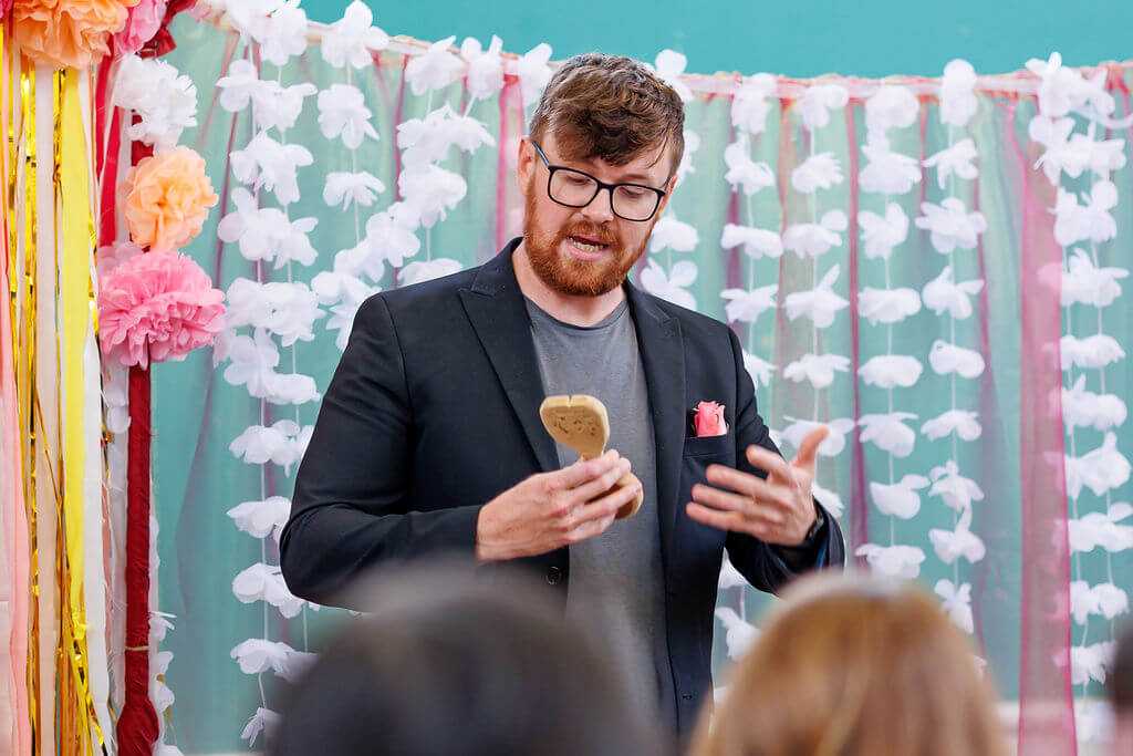 A man with a beard, wearing glasses and a black blazer, is reading a message from the sole of a shoe. He's standing in front of a decorative backdrop adorned for the wedding ceremony with colorful flowers and white ribbons. Guests are seated attentively in front of him.