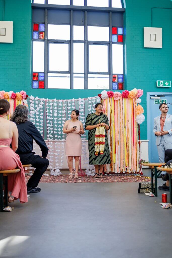 People are gathered in a colorful room with a decorative backdrop of streamers, flowers, and a garland. At the heart of this vibrant setting, two individuals stand, one holding a microphone. Stained glass windows glow above as attendees sit engaged with the speakers at what feels like a wedding ceremony.