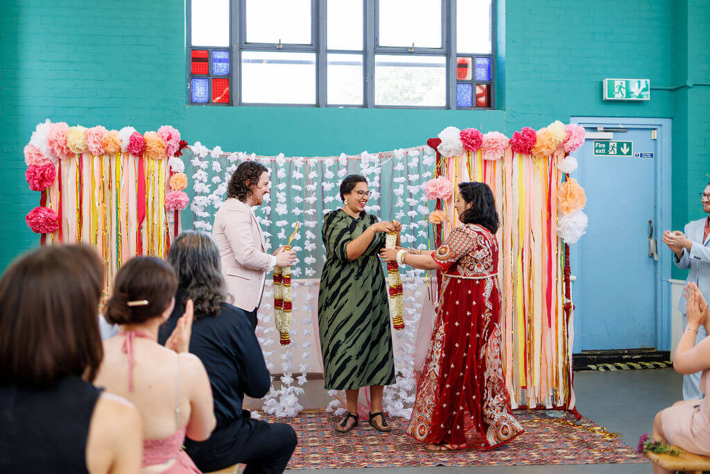 A joyful indoor wedding ceremony unfolds with three people standing at the front. They incorporate a garland exchange in a decorated area with colorful flowers and ribbons. Attendees watch and clap as sunlight pours through the large window, illuminating the teal wall.