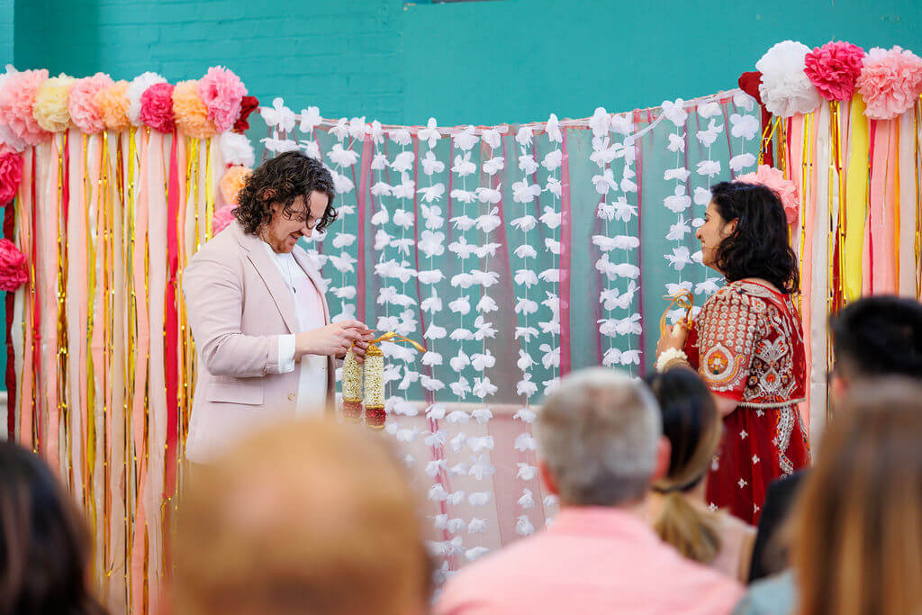 In a vibrant wedding ceremony setting, a couple stands facing each other. The person on the left is in a light suit, and the person on the right dazzles in red and gold. They are incorporating tradition with a garland exchange against a backdrop adorned with hanging flowers.