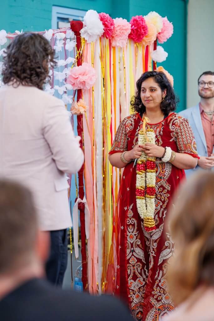 A woman in a red and gold embroidered dress stands holding a flower garland, smiling at a man in a light pink blazer. They are participating in the Garland Exchange during the wedding ceremony, framed by a colorful backdrop with paper flowers and ribbons. Another man in a blue suit smiles from the background.