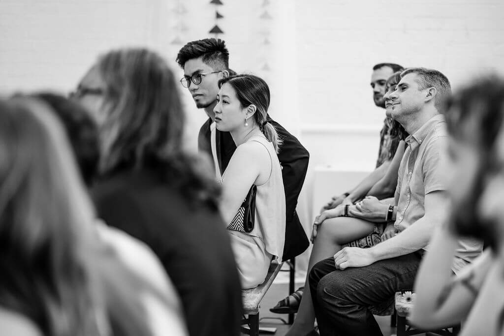 In a black and white photo, a seated group of people attentively listens, with two individuals at the center of focus. The room, minimally decorated, suggests an intimate setting—perhaps capturing a special moment during a wedding ceremony.