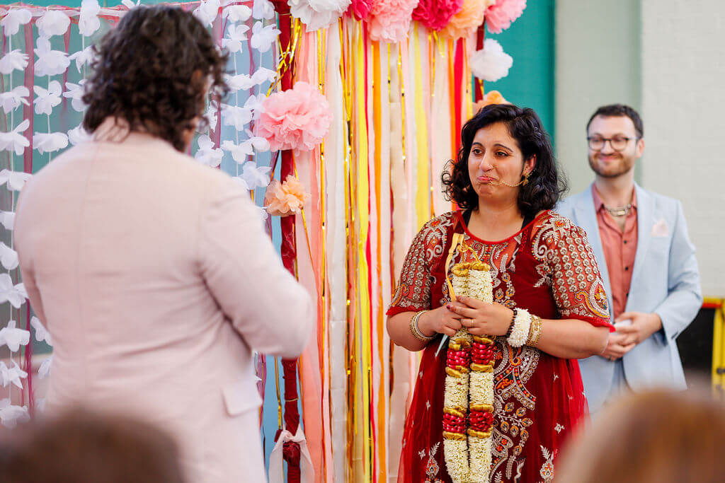 At their wedding ceremony, a woman in a red and gold dress holds a floral garland, poised for the garland exchange with a man in a light suit. They stand amid vibrant floral decorations, while the smiling officiant presides nearby.