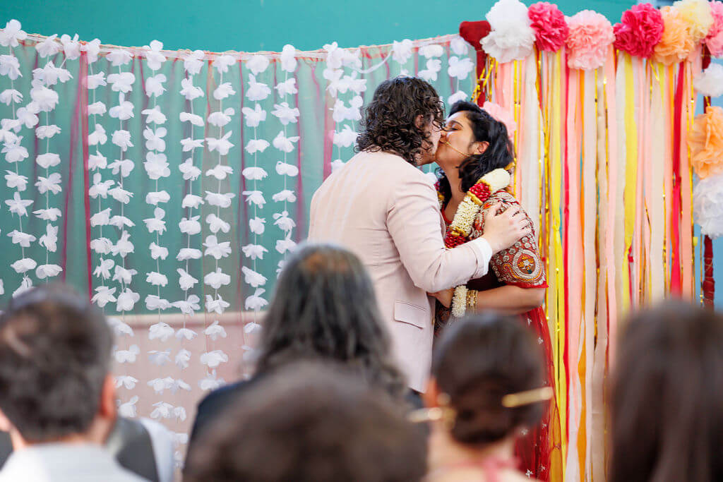 A couple shares a kiss in front of a vibrant, floral backdrop adorned with paper flowers and streamers during their wedding ceremony. As guests seated in chairs witness the garland exchange, the atmosphere feels festive and celebratory.