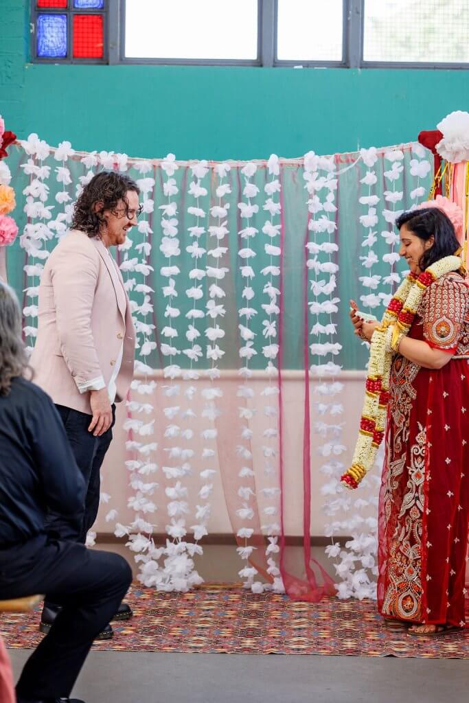 A couple stands in front of a floral curtain backdrop during their wedding ceremony. The woman, in a red and gold traditional outfit, holds a garland ready for the garland exchange. The man, wearing a light suit, faces her as a seated guest watches the scene unfold.