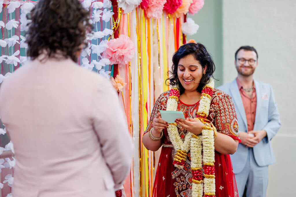 At a vibrant wedding ceremony, a woman in a red traditional dress and floral garlands reads from a card, smiling. She stands beside a backdrop of pink and white flowers. In the background, during the garland exchange ritual, a man in a suit beams with delight.