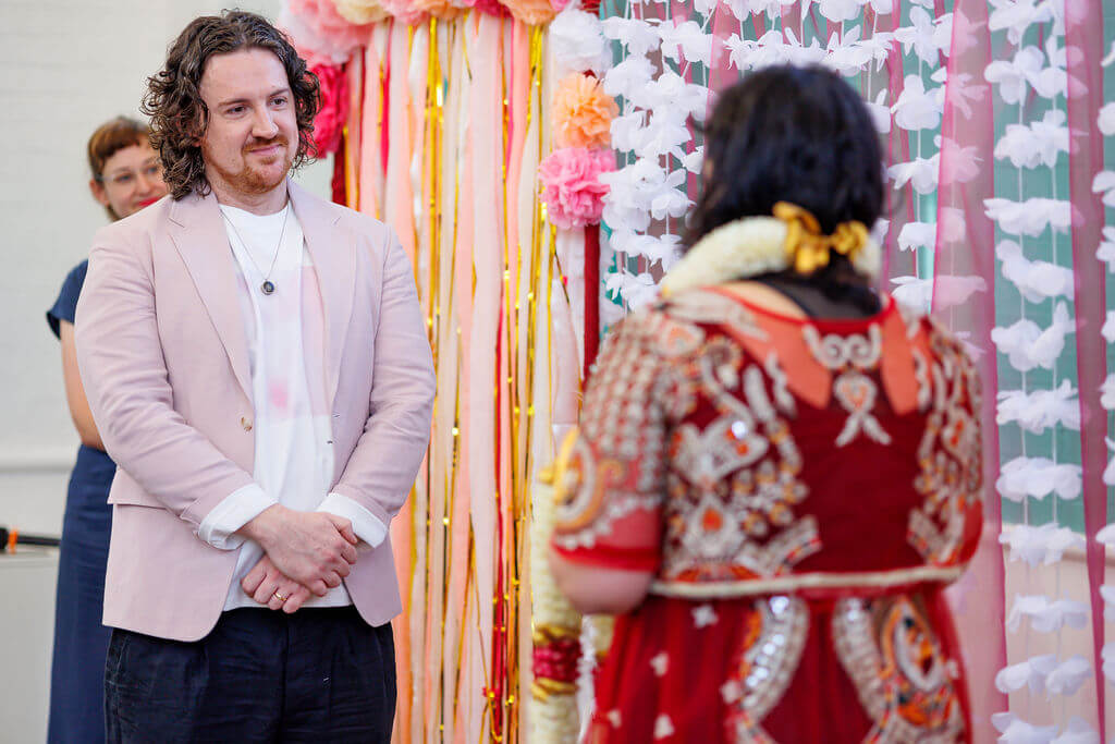 A person in a pastel pink blazer and white shirt smiles during the garland exchange at a wedding ceremony. The other individual, in an ornate red and gold outfit adorned with floral garlands, stands before a colorful backdrop of pink and white flowers interwoven with gold streamers.