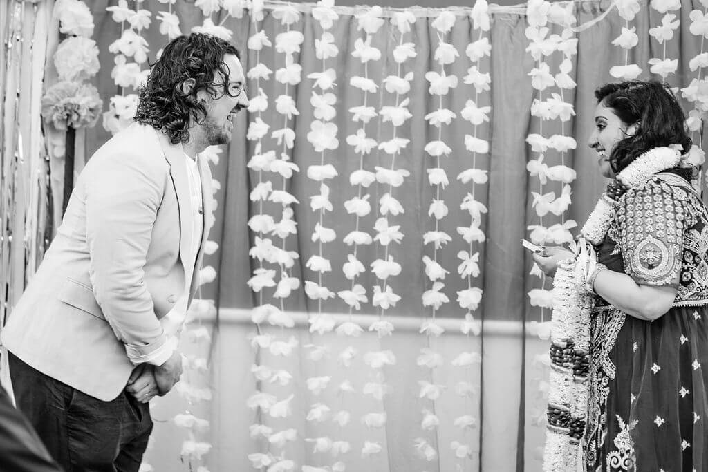 A black-and-white photo captures two people smiling at each other in front of a floral backdrop at what appears to be a wedding ceremony. One wears a suit, the other is in traditional attire adorned with floral garlands, suggesting a heartfelt garland exchange.