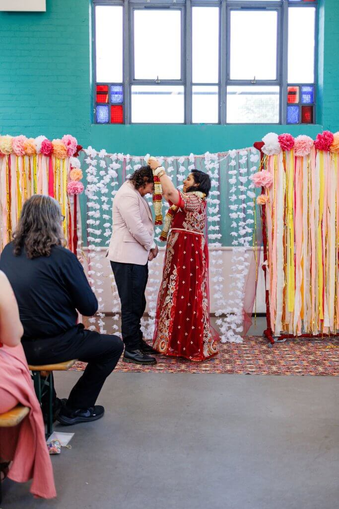 A couple stands in front of a colorful backdrop during their wedding ceremony. The woman, in a red and gold outfit, performs the garland exchange, placing it gracefully over the man's neck. Decorative strings and paper flowers adorn the scene, with guests seated nearby.