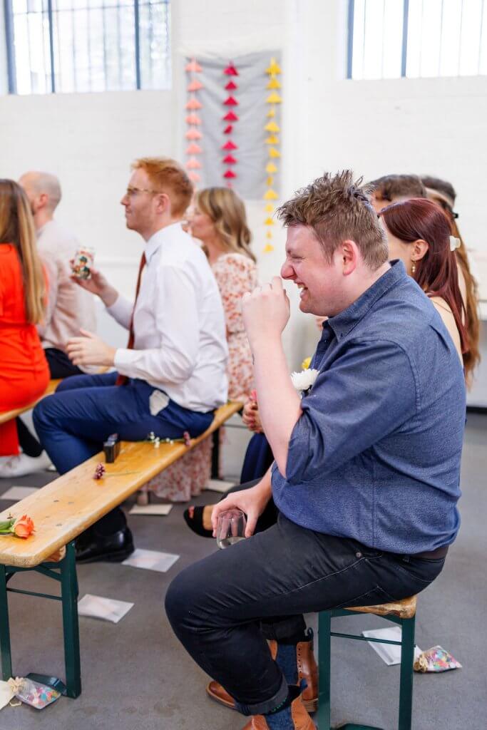 A group of people are seated on wooden benches in a bright room with white walls and a colorful paper crane decoration. The focus is on a man in a blue shirt laughing while holding a glass, as the joyful atmosphere hints at an upcoming wedding ceremony.
