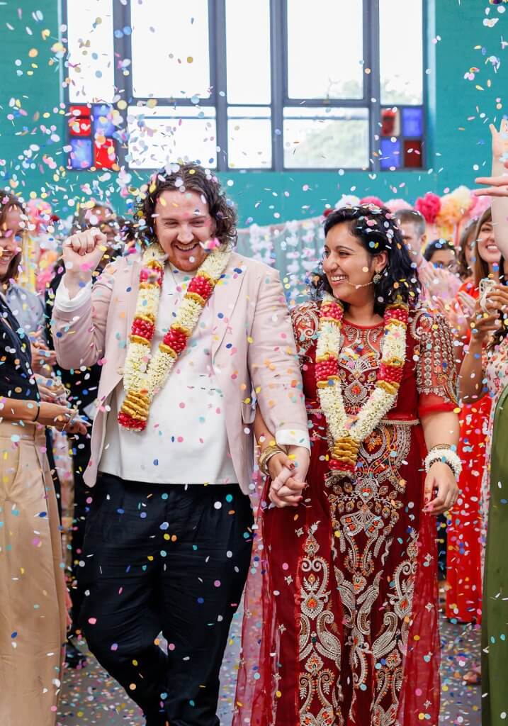 A couple, fresh from their garland exchange, wearing traditional wedding attire, joyfully walks hand-in-hand through a shower of colorful confetti in a bright room. They are surrounded by cheering guests, celebrating their union at this beautiful wedding ceremony.