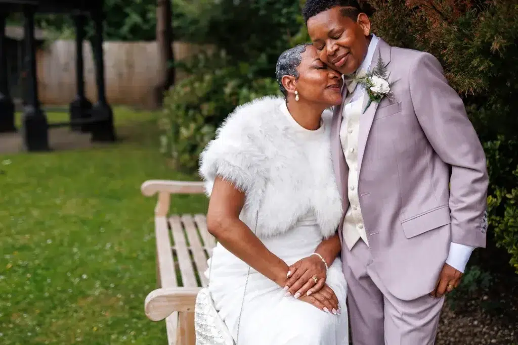 Gentle Couples Portrait of two brides in the gardens of The Clarendon Hotel in Blackheath. One is in a white mermaid dress with a faux fur coat leaning into the other bride standing in a pink suit.
