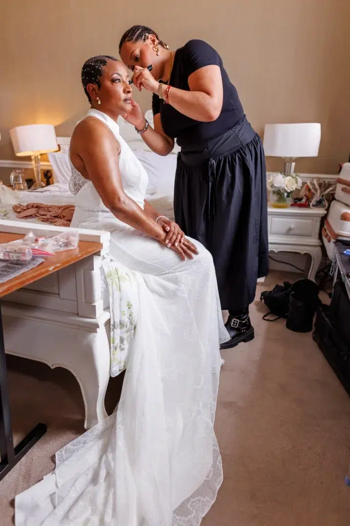 LGBTQ+ Wedding Getting Ready at The Clarendon Hotel - Black Woman in Long White Wedding Dress sitting on the bed and hair and makeup person is styling short hair with pearls